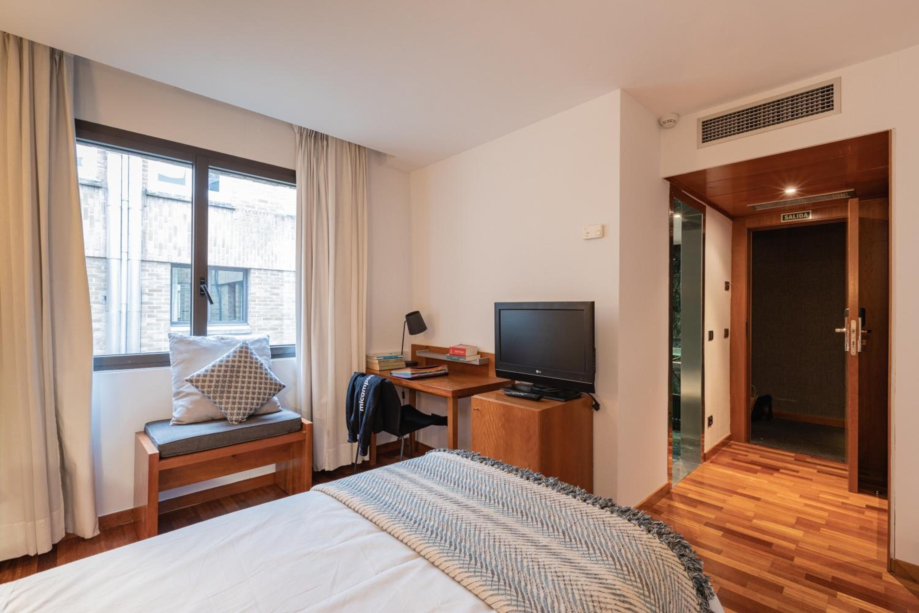 Une chambre d’hôtel avec un lit, un bureau avec une télévision, une fenêtre avec des rideaux et une porte portant l’inscription « SORTIE ».