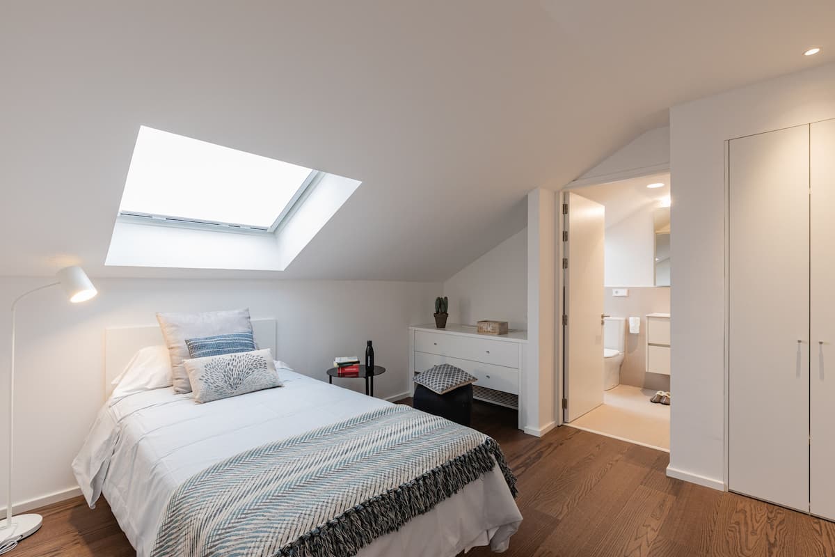 Une chambre avec une lucarne, un lit recouvert d’une couvre-lit à motifs, une table de chevet avec une lampe, et une vue sur la salle de bain.