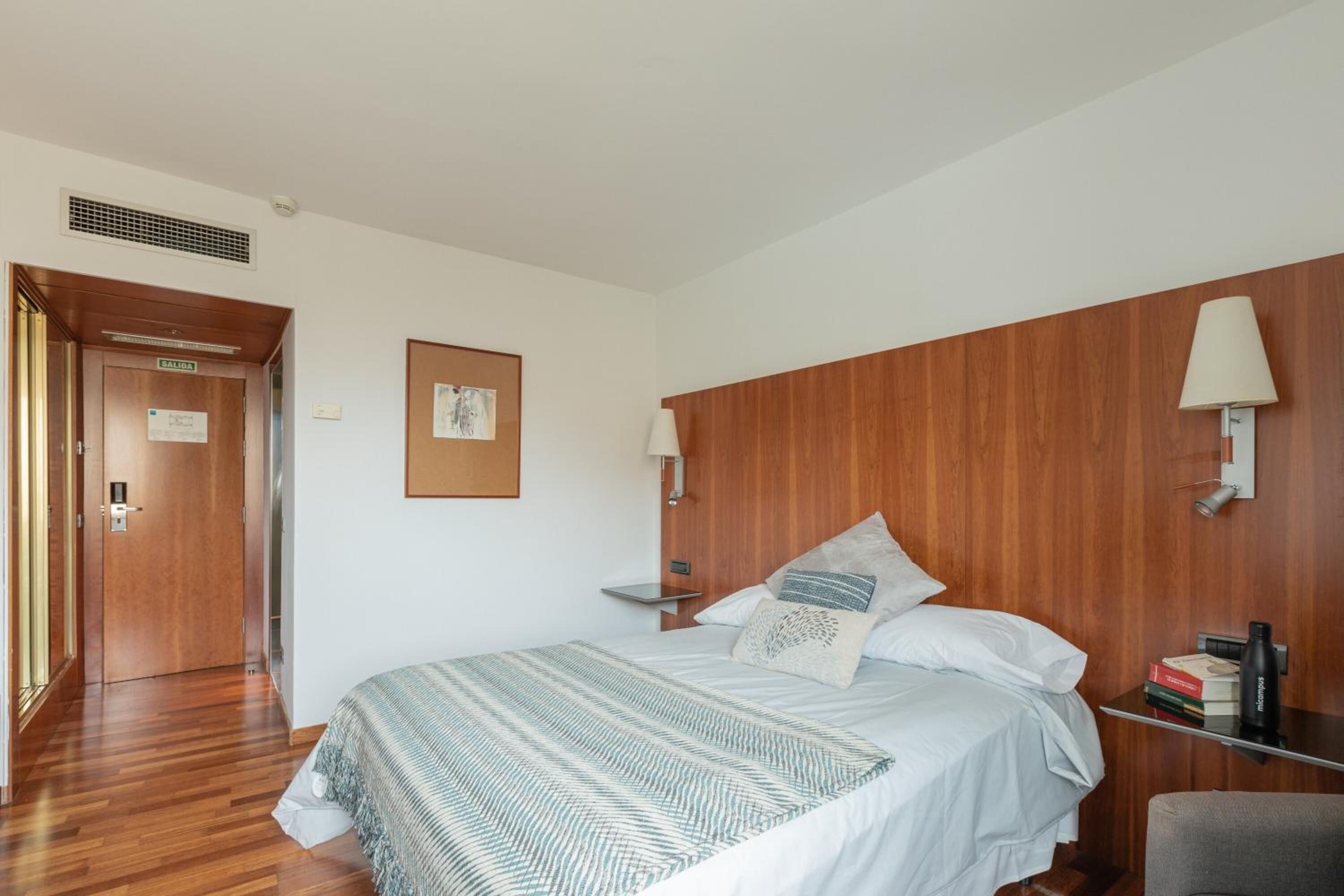 Une chambre d’hôtel avec une tête de lit en bois, un lit recouvert d’une couverture à rayures, une table de chevet avec une lampe et une petite étagère avec des livres et une bouteille d’eau.
