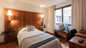A hotel room with a wooden headboard, a bed with a striped blanket, bedside tables with lamps, a window with curtains, and a desk with a remote control. - Micampus Pamplona  (Pamplona)