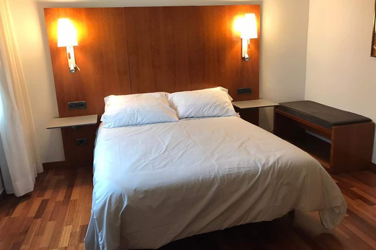 Une chambre d’hôtel avec une tête de lit en bois, deux lampes de chevet, un lit avec du linge de lit blanc, une table de chevet et un bureau avec une chaise.