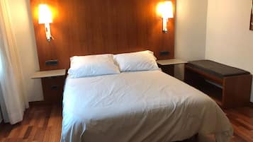 Quarto de hotel com cabeceira de madeira, duas luminárias de cabeceira, cama com roupa de cama branca, mesa de cabeceira e escrivaninha com cadeira.