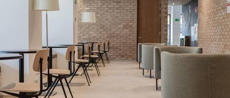 A modern hotel lobby with a brick wall, a reception desk, and a row of chairs and tables.