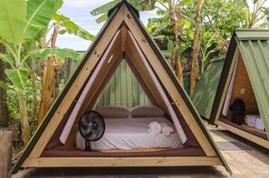 A triangular wooden tent with a bed, pillows, and a fan. - ClanDestino Hostel (Ilhabela)