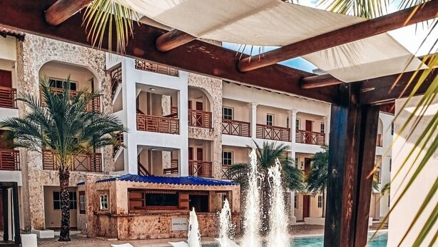 Área de piscina de hotel com uma fonte, palmeiras e um edifício com várias andares e sacadas.