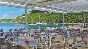 Een terras met tafels en stoelen, met uitzicht op een strand met parasols en ligbedden.
