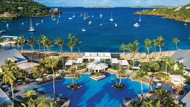 Een resort met een centraal gelegen zwembad, palmbomen en uitzicht op zee met boten.