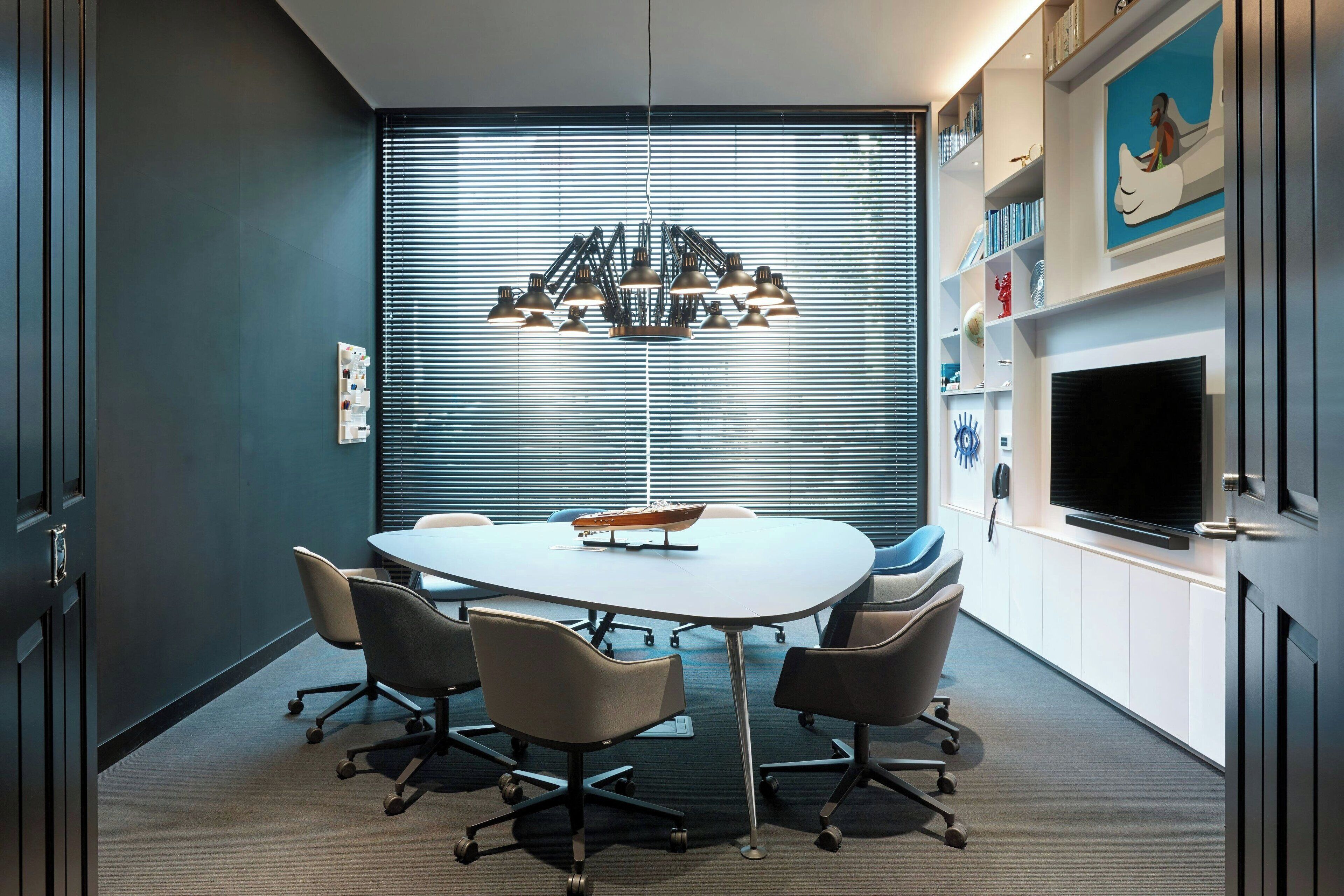 A modern meeting room with a round table, four chairs, a chandelier, a TV, and a bookshelf with decorative items.