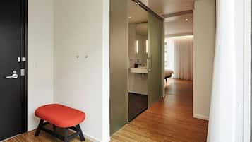 Una habitación de hotel moderna con suelo de madera, un banco rojo, una puerta corredera de vidrio hacia un baño y un ventanal con cortinas.