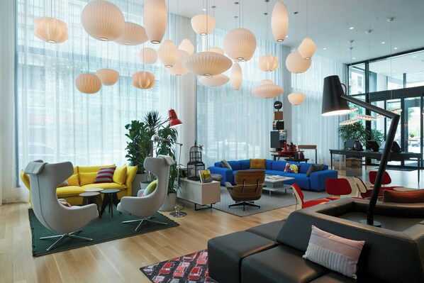 A modern living room with a yellow sofa, a blue sofa, and a black sofa. There are multiple hanging lights and a large window in the background. - citizenM Los Angeles Downtown (Los Angeles)