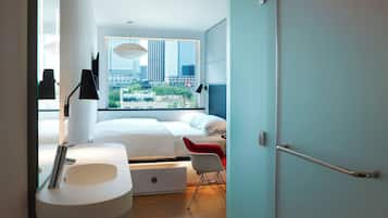 A modern bedroom with a large bed, a bedside table, a red chair, and a view of a cityscape through a large window.
