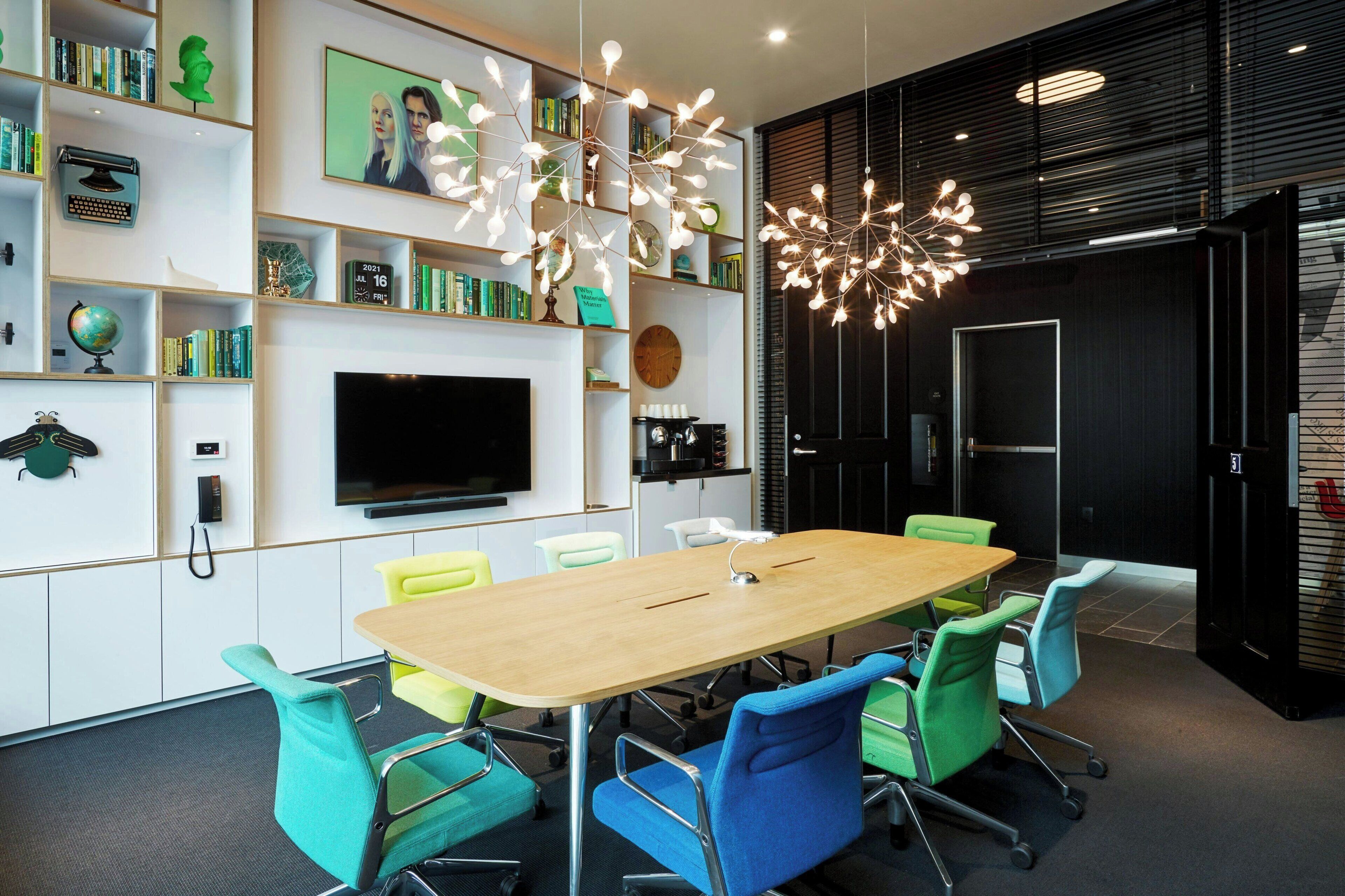 Een moderne vergaderruimte met een lange tafel, kleurrijke stoelen, een grote flatscreen-tv en een decoratieve lamp.