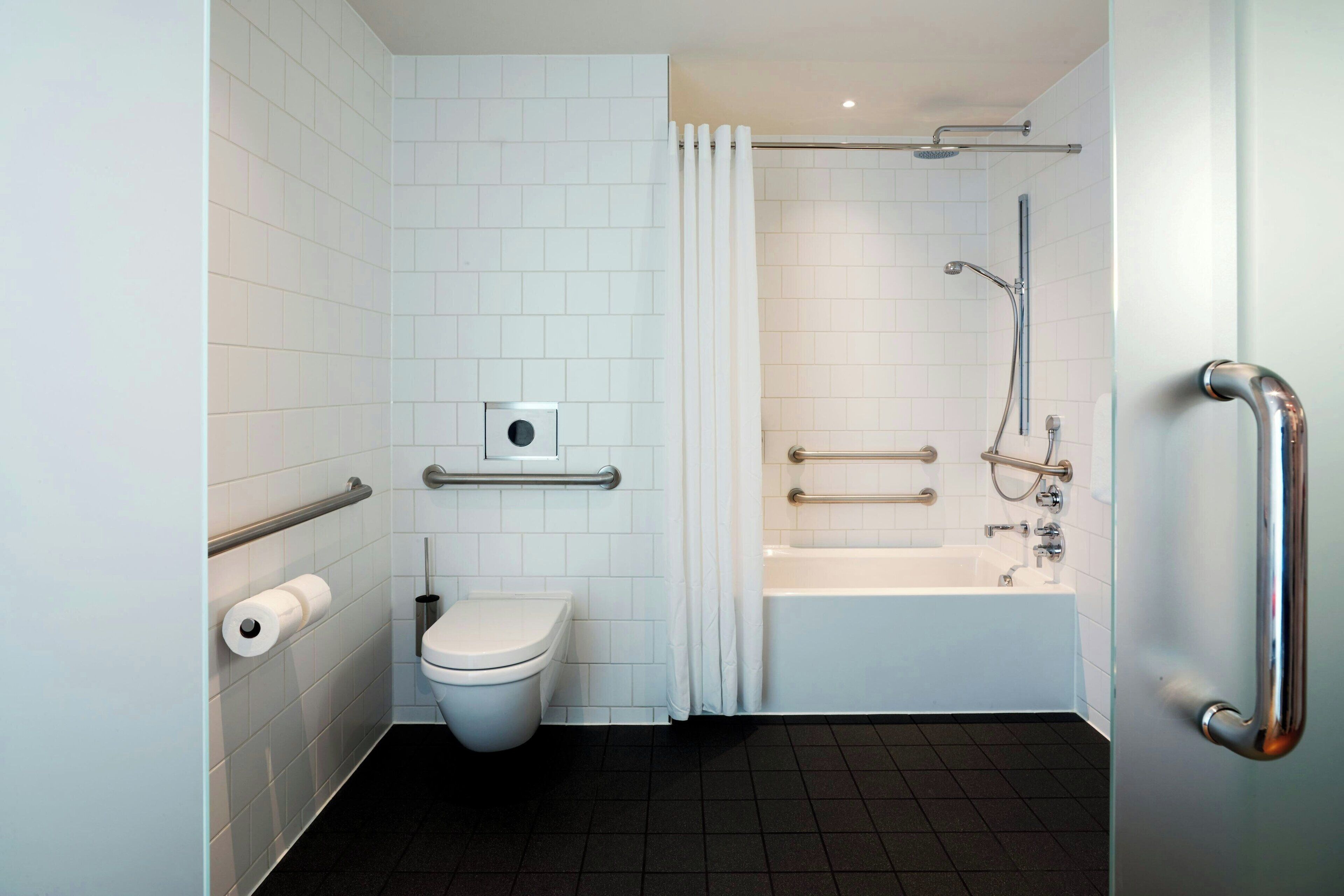 Een moderne badkamer met witte tegels, een toilet, een bad met handgrepen en een douchehoek.