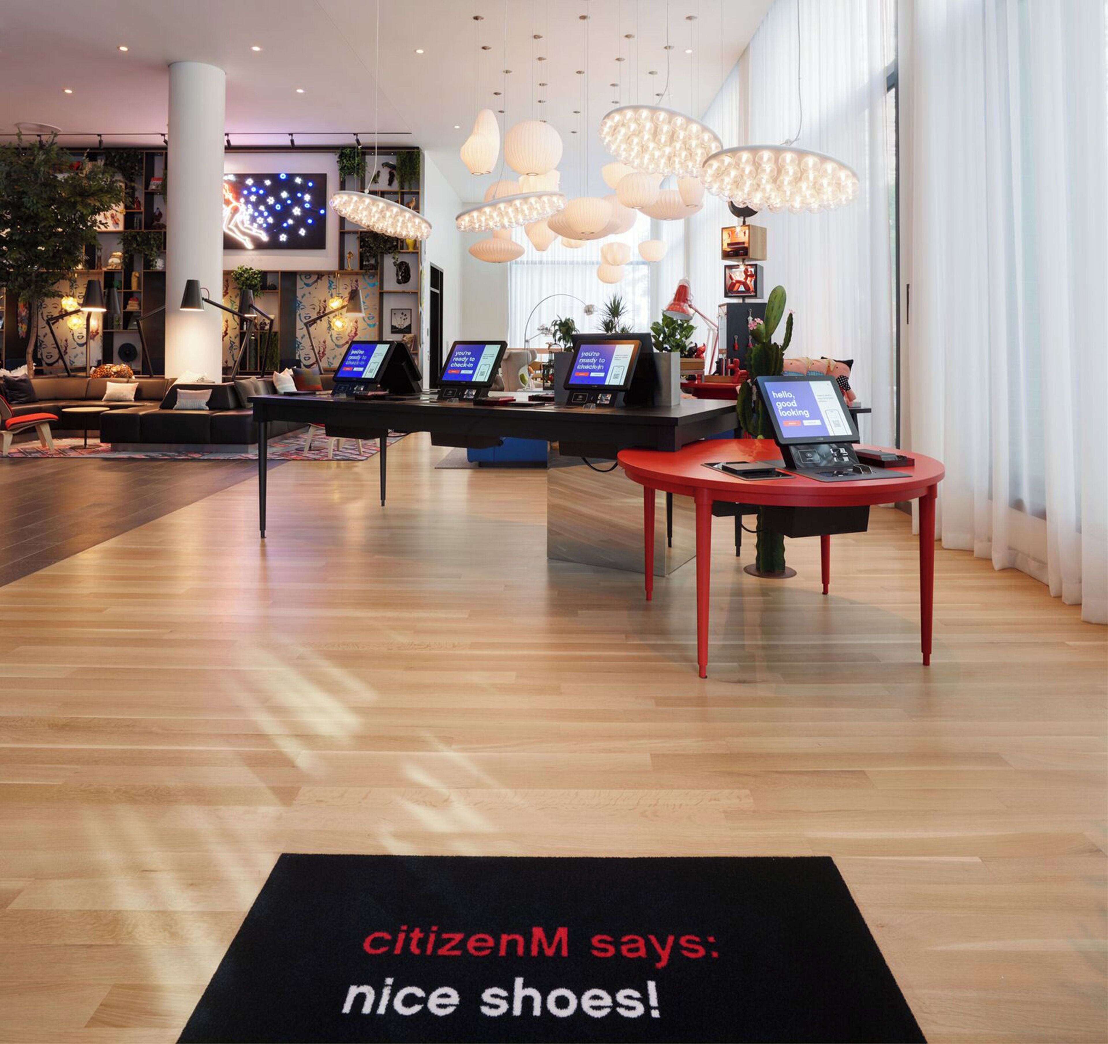 A modern lobby with a reception desk, digital check-in kiosks, and a red table with a laptop. A black rug with 'citizenM says: nice shoes!' is visible.