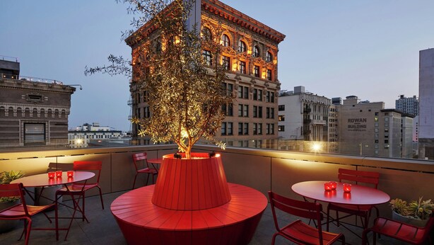 A rooftop terrace with red seating and a central tree, overlooking a cityscape with historic buildings.