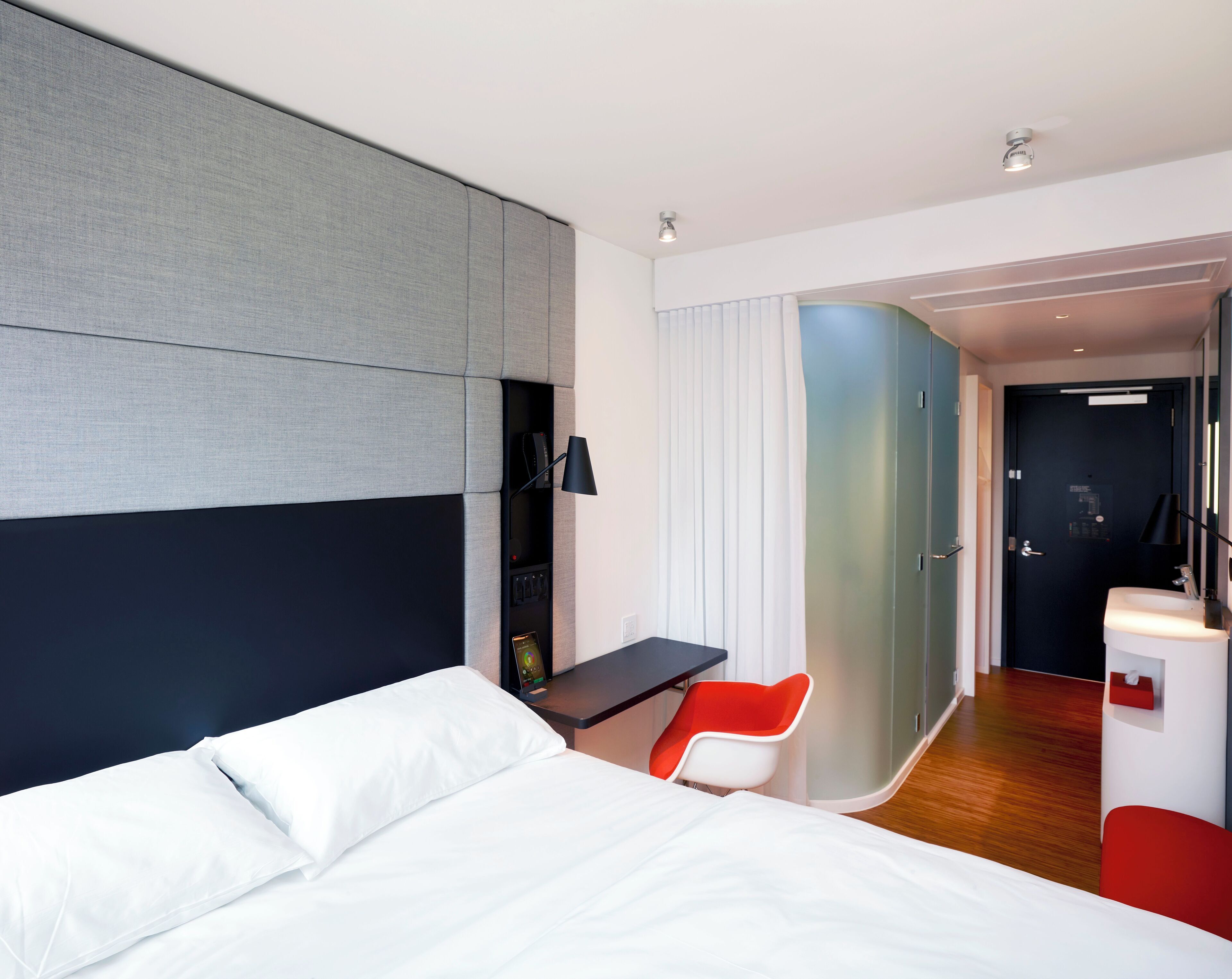 A modern hotel room with a large bed, a desk, a red chair, and a bathroom with a mirror and sink.