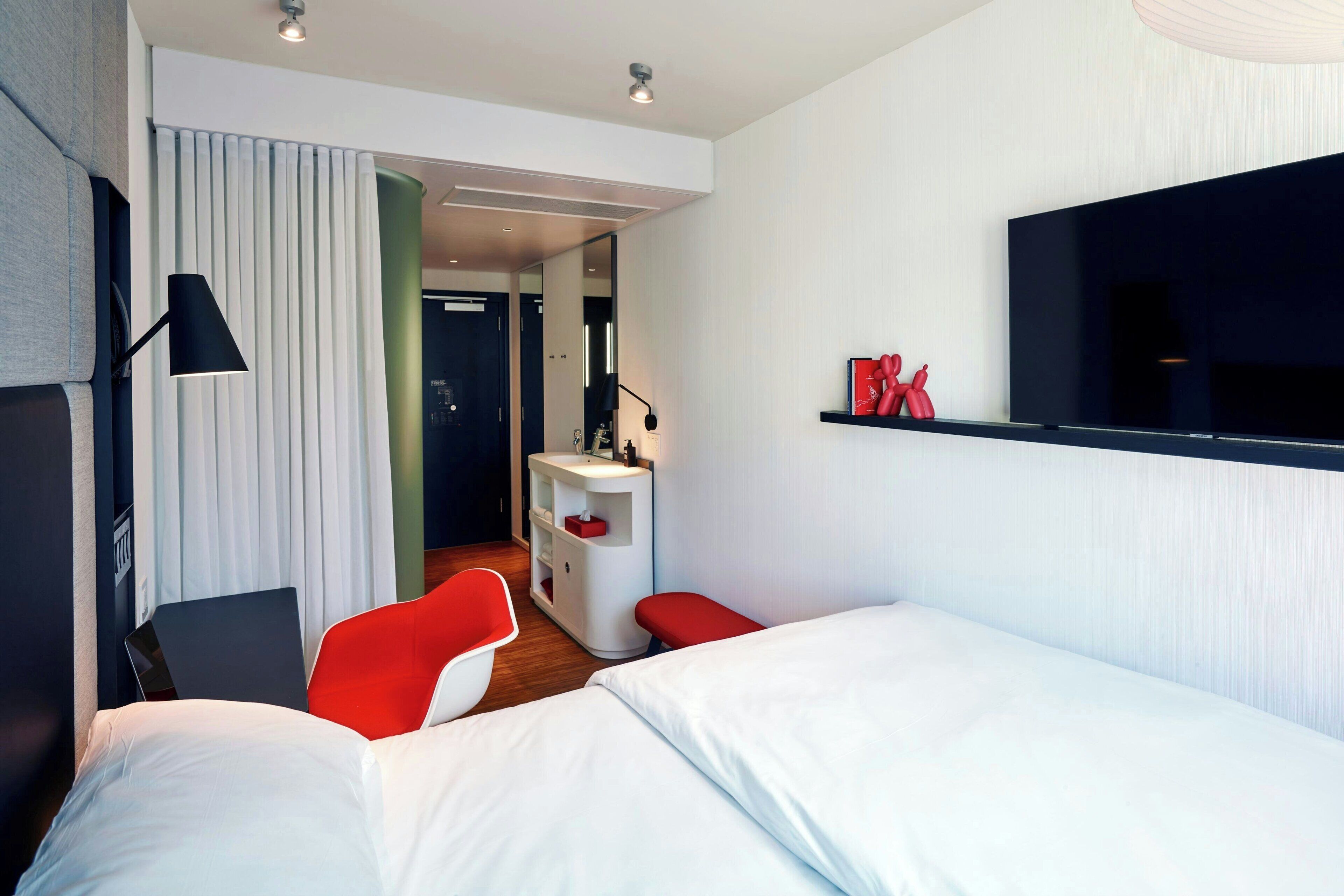 A modern hotel room with a bed, a desk, a chair, a vanity, and a TV.