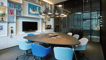 Une salle de conférence dotée d’une grande table, de chaises bleues, d’un téléviseur fixé au mur et d’étagères remplies de livres et d’objets décoratifs.