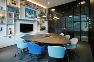 A conference room with a large table, blue chairs, a mounted TV, and shelves with books and decorative items.
