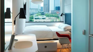 A modern hotel room with a large bed, a desk, a chair, a round sink, and a window with a city view.