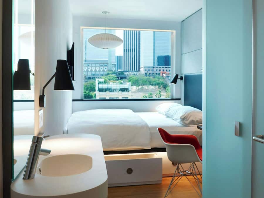 A modern hotel room with a large bed, a desk, a chair, a round sink, and a window with a city view.