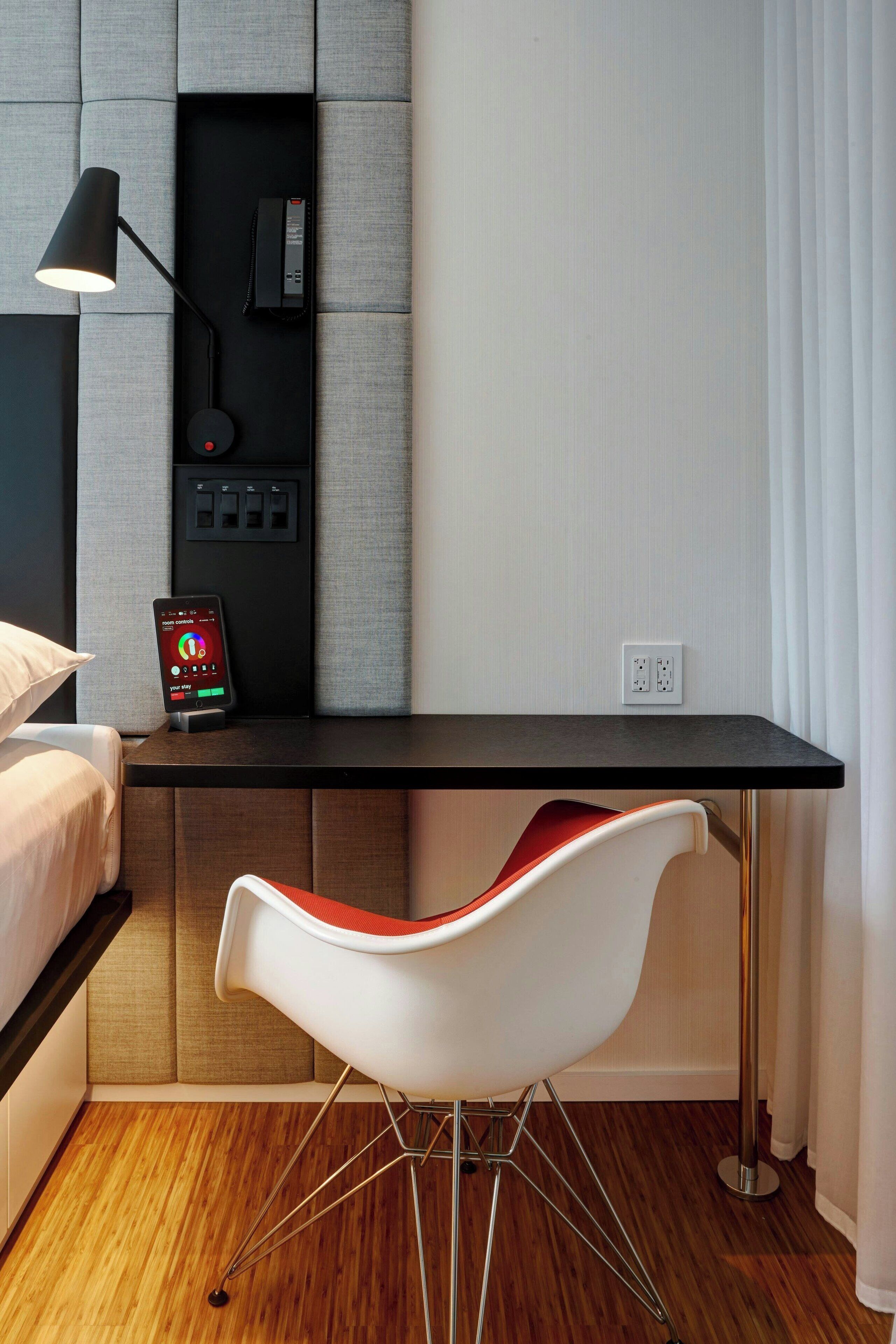 A modern hotel room with a wooden floor, a black desk with a smartphone on it, a white chair, and a bed with a white pillow.