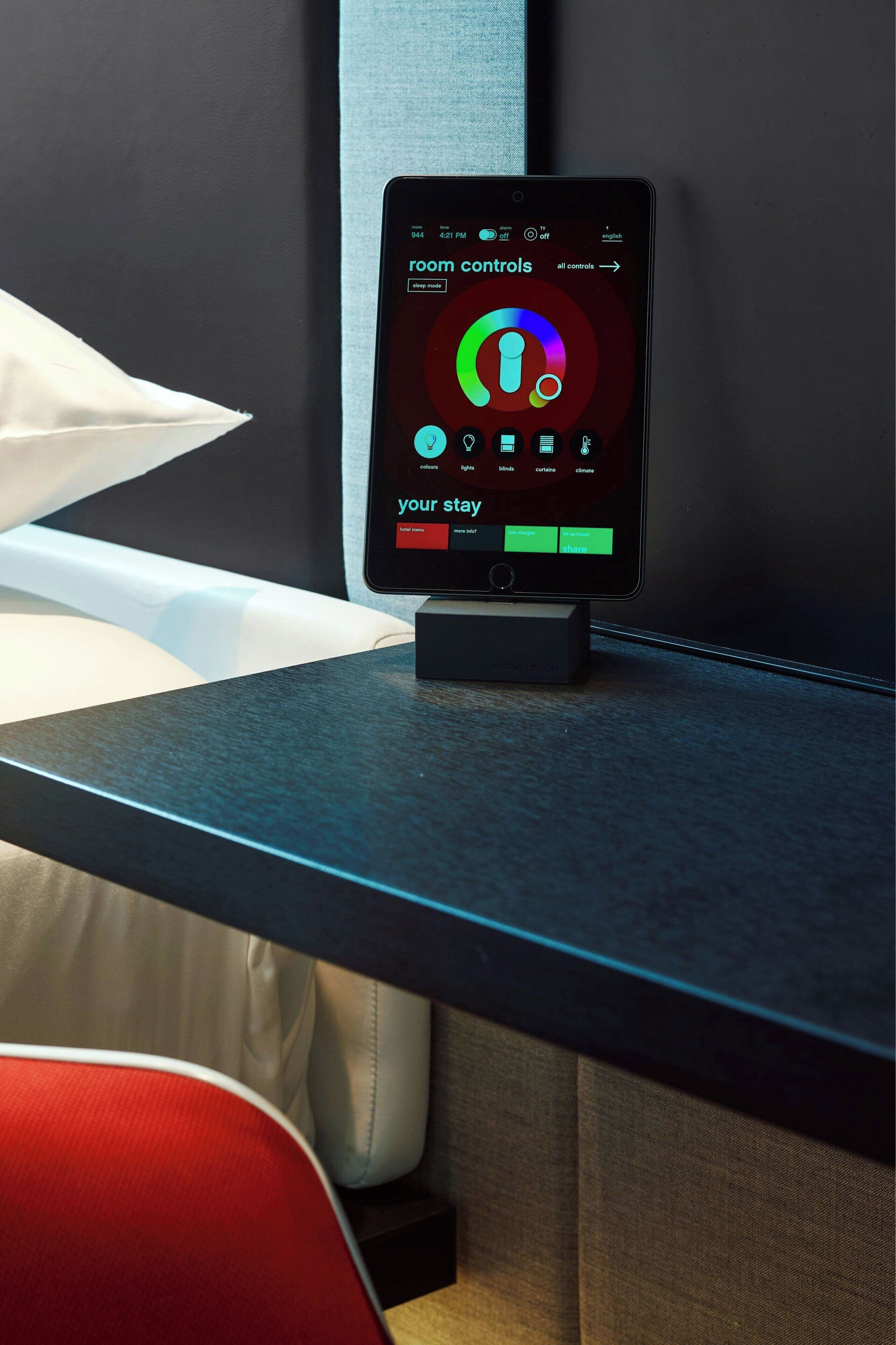A smartphone on a charging dock displaying a room control app with options for lighting, blinds, and climate control.