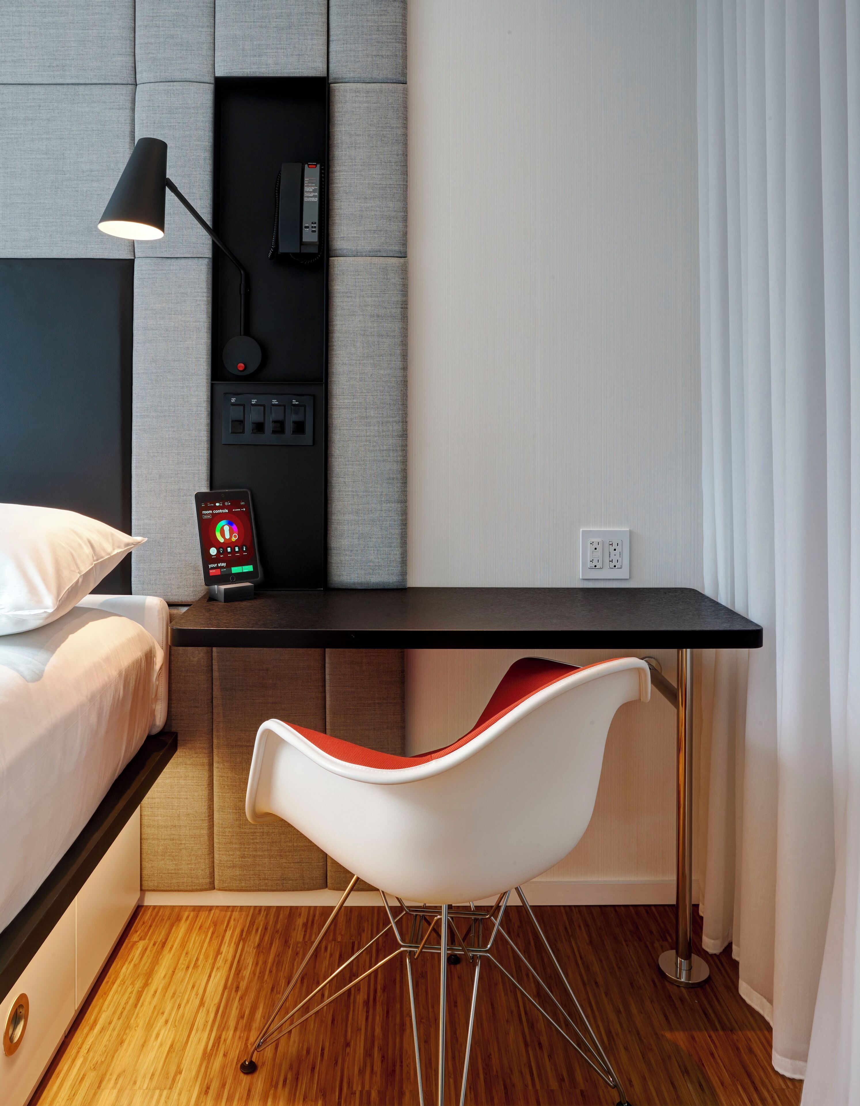 A modern hotel room with a bed, a desk, a chair, and a bedside table.