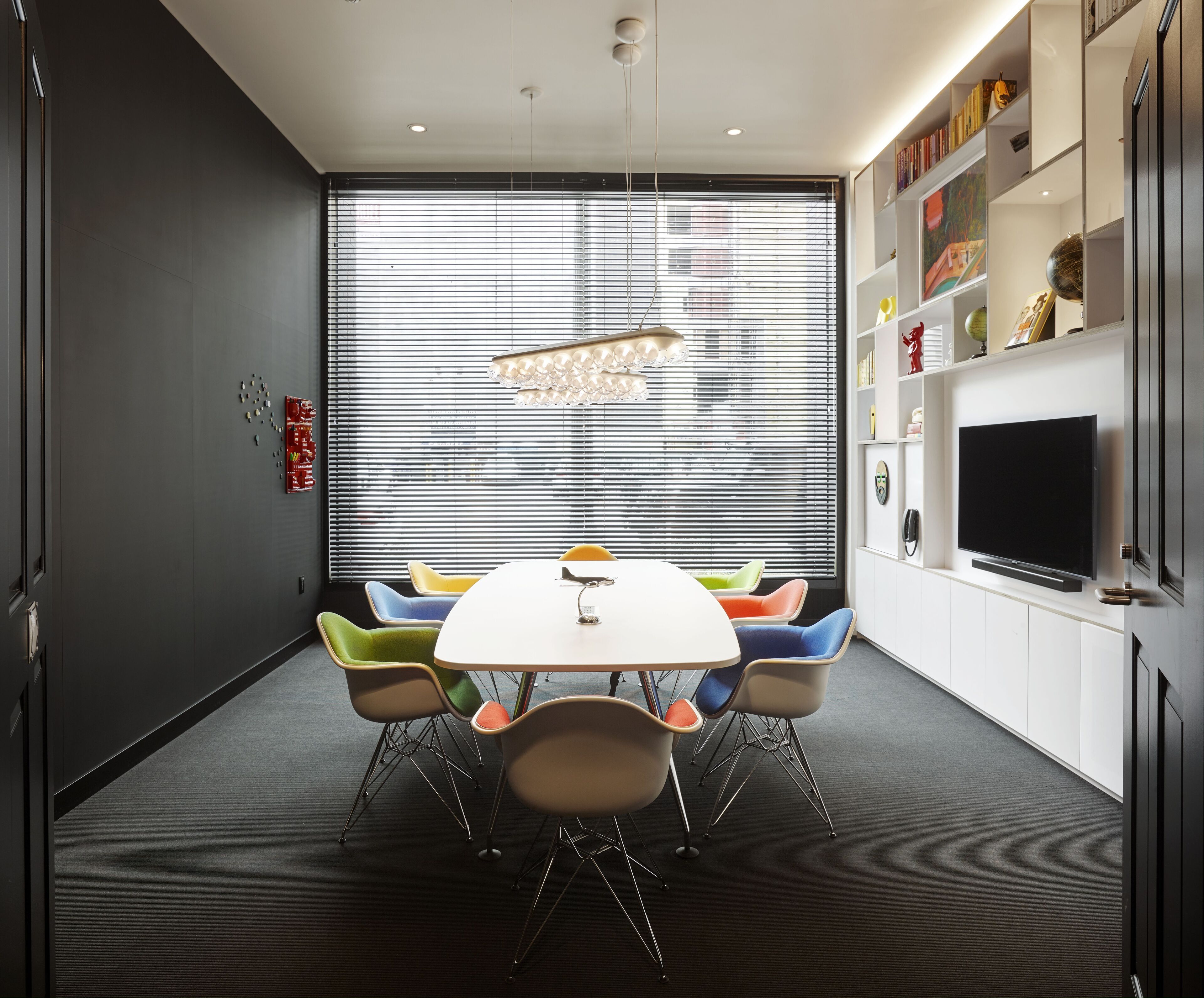 A modern meeting room with a white table, colorful chairs, a chandelier, and a TV on a white cabinet.