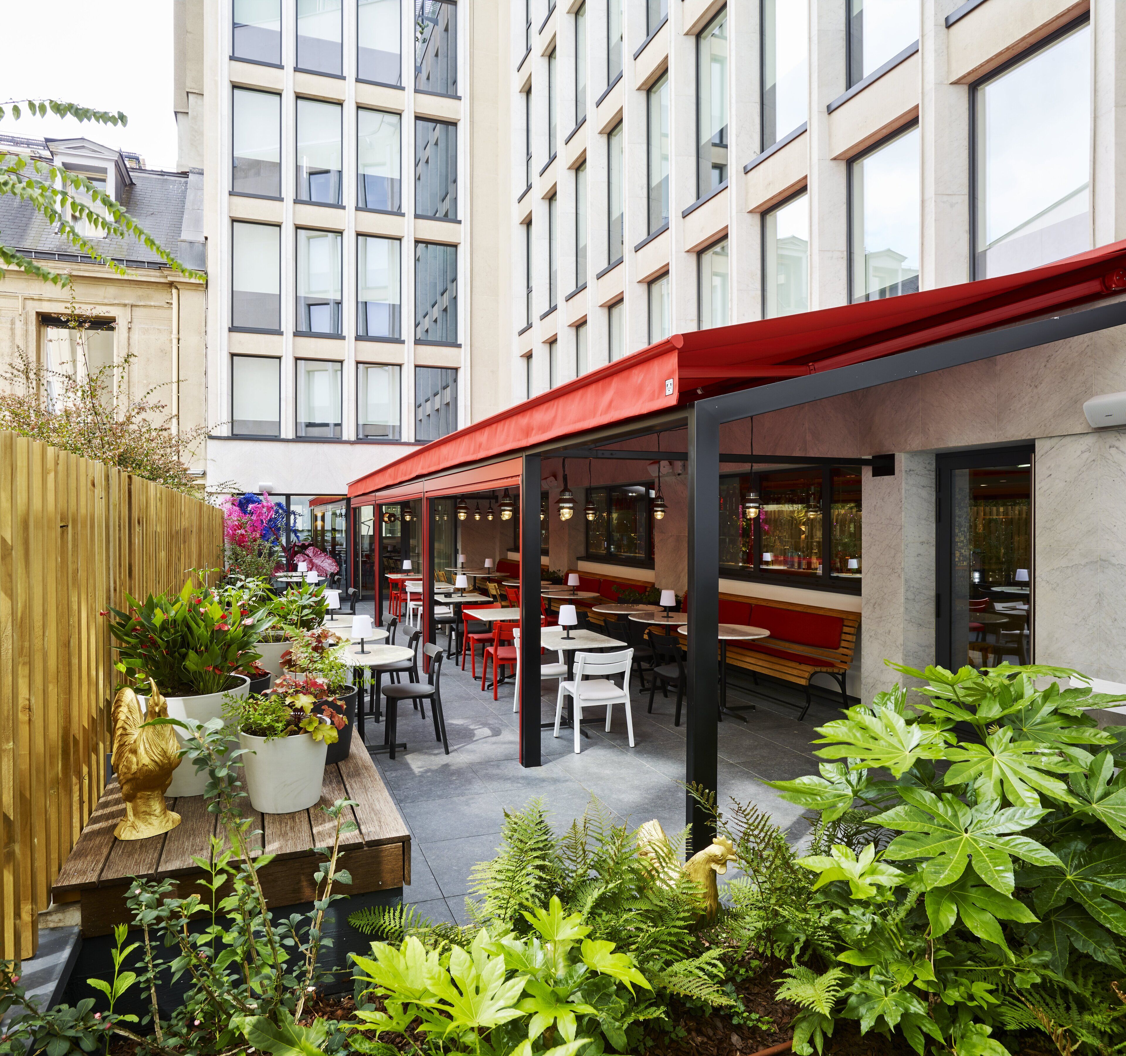 An outdoor café with tables and chairs, surrounded by potted plants and greenery.