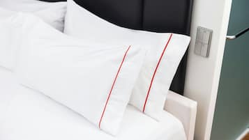 A neatly made bed with white bedding and red trim, a black headboard, and a wall-mounted light switch.