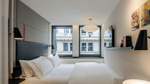 A modern hotel room with a large bed, a desk, a chair, and a view of a building through a large window. - citizenM Paris Champs-Élysées (Paris)