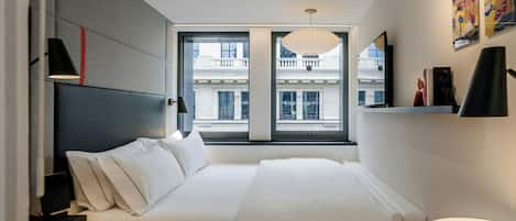 Habitación de hotel moderna con una cama grande, un escritorio, una silla y vistas a un edificio a través de un ventanal amplio.