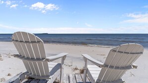 Deux chaises Adirondack blanches sont placées sur une terrasse en bois, face à une plage où la mer est calme et le ciel dégagé.