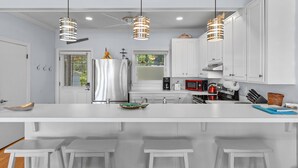 Une cuisine moderne avec des meubles blancs, un grand îlot central doté de tabourets, des luminaires suspendus et un réfrigérateur en acier inoxydable.