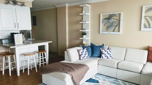 A living room with a white sofa, a coffee table, a kitchen area with a bar, and a built-in shelving unit. - Condo right on the beach! Stunning seventh floor views! (Galveston)