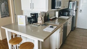 A kitchen with white cabinets, a microwave, a refrigerator, a coffee maker, and a sink. There are two wooden stools at the kitchen island. - Condo right on the beach! Stunning seventh floor views! (Galveston)