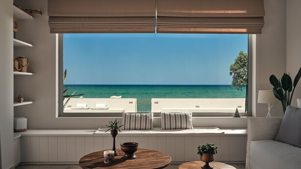 Una sala moderna con un ventanal que da a una playa, un sofá, dos mesas de madera y una consola blanca con adornos.