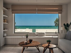 A modern living room with a large window overlooking a beach, a sofa, two wooden tables, and a white console table with decorative items. - Astarte Villas - Onda Del Mar Beach Villa (QW98+2V2 Argassi, Greece)