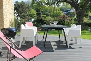 Een terras buiten met een tafel en witte stoelen, een barbecue en een rode lounge stoel.