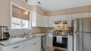 A modern kitchen with white cabinets, stainless steel appliances, a coffee maker, and a window with blinds. - Postcards from the Bay (Norfolk)