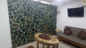 Una sala de estar con pared de piedra, un sofá con cojines, un taburete de madera y un televisor montado en la pared.