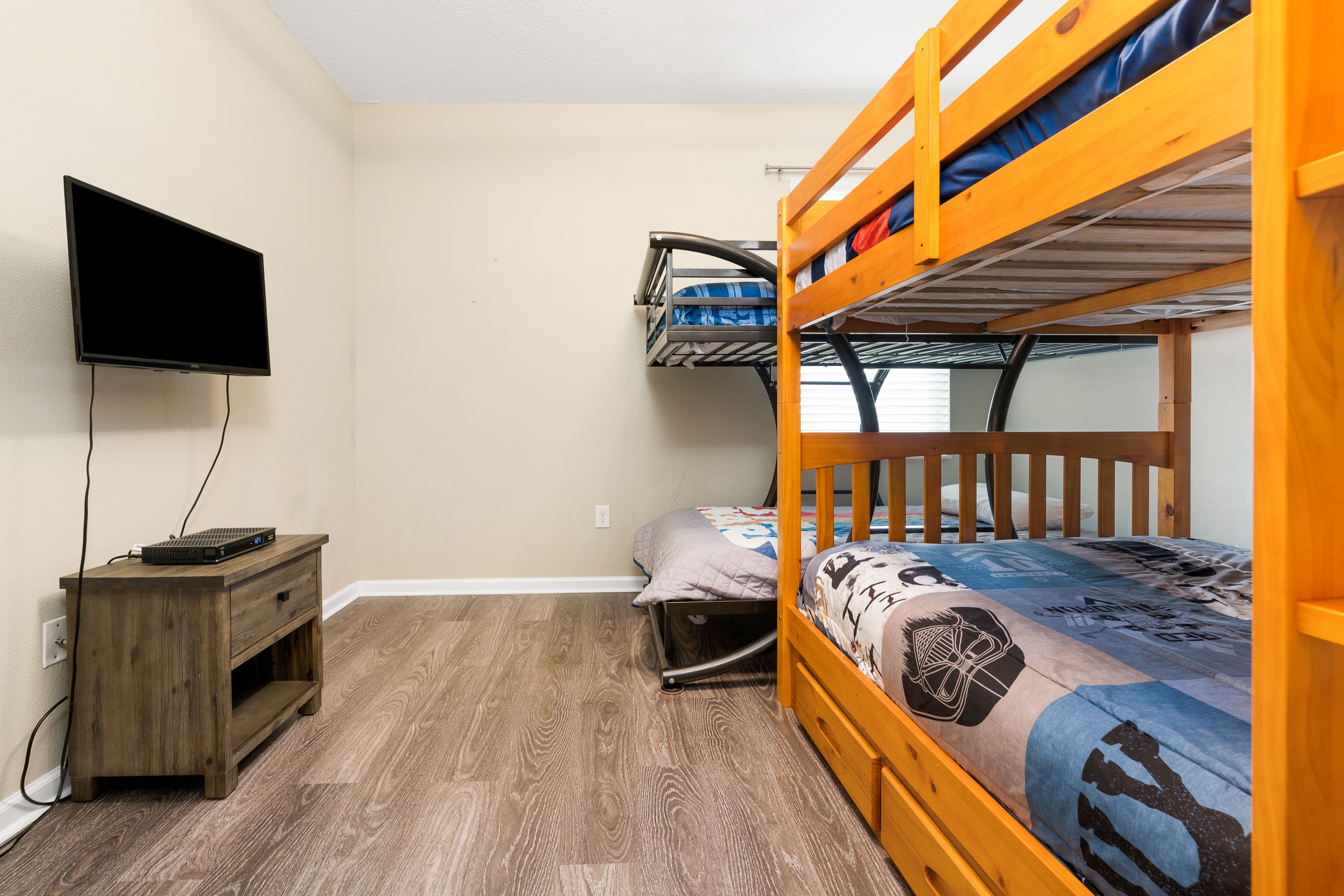 A bunk bed room with a TV on a wooden stand, a small wooden table, and a window with blinds.