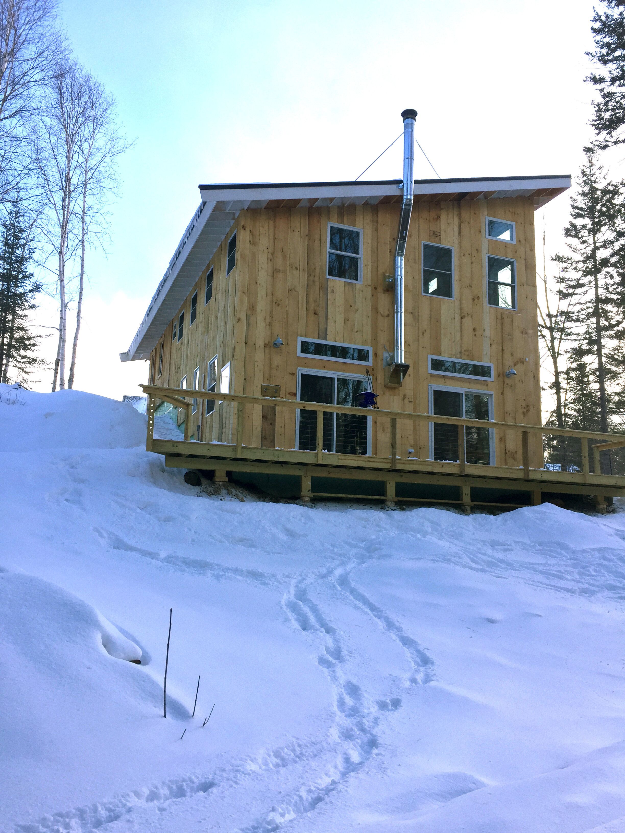 A wooden cabin with multiple windows and a chimney, surrounded by snow and trees.