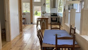 Ett rymligt matområde med ett träbord och stolar, en kristallkrona och utsikt över köket och trädgården genom stora fönster.
