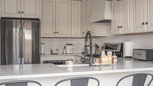 Een moderne keuken met witte kasten, roestvrijstalen apparatuur en een marmeren aanrecht. Drie barkrukken met houten zitplaatsen.