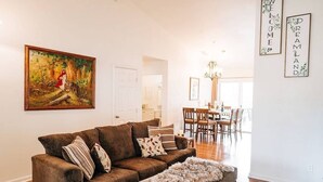 A living room with a brown sofa, a fur throw, and a painting of a woman in a forest. There is a dining area with wooden chairs and a chandelier. - Views of Bass Lake, whole house generator, BBQ, Outdoor dining, Mountain Views  (Madera County)