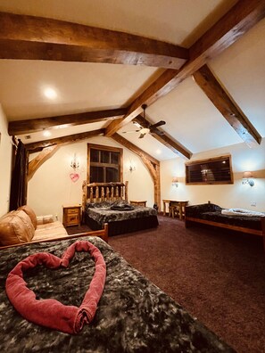 A bedroom with a vaulted ceiling, wooden beams, a ceiling fan, two beds, a window with blinds, and a chandelier.
