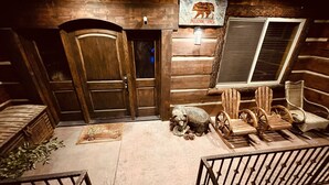 A log cabin interior with wooden doors, a bear sculpture, Adirondack chairs, and a window with blinds.