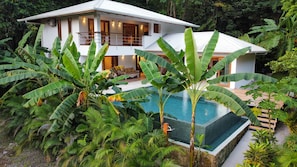 A two-story house with a swimming pool surrounded by tropical plants.